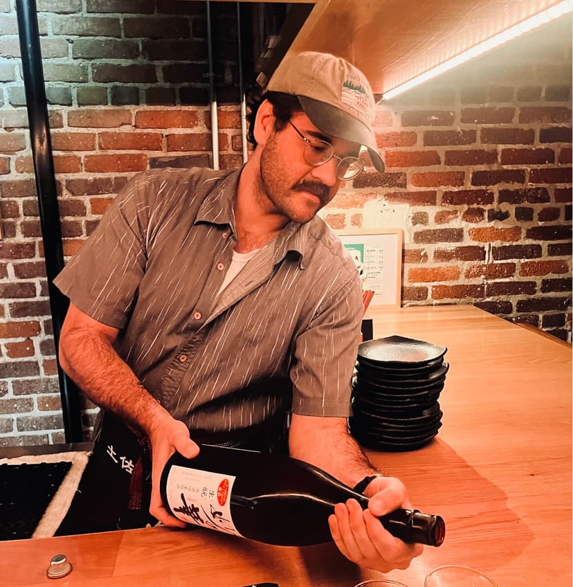 Photo of me pouring sake from a large bottle at a bar. The bar is wood. Behind me is an exposed brick wall. I'm a man in my 30s with a mustache and glasses. I'm wearing a beige baseball cap, glasses, and a grey short-sleeve button down.